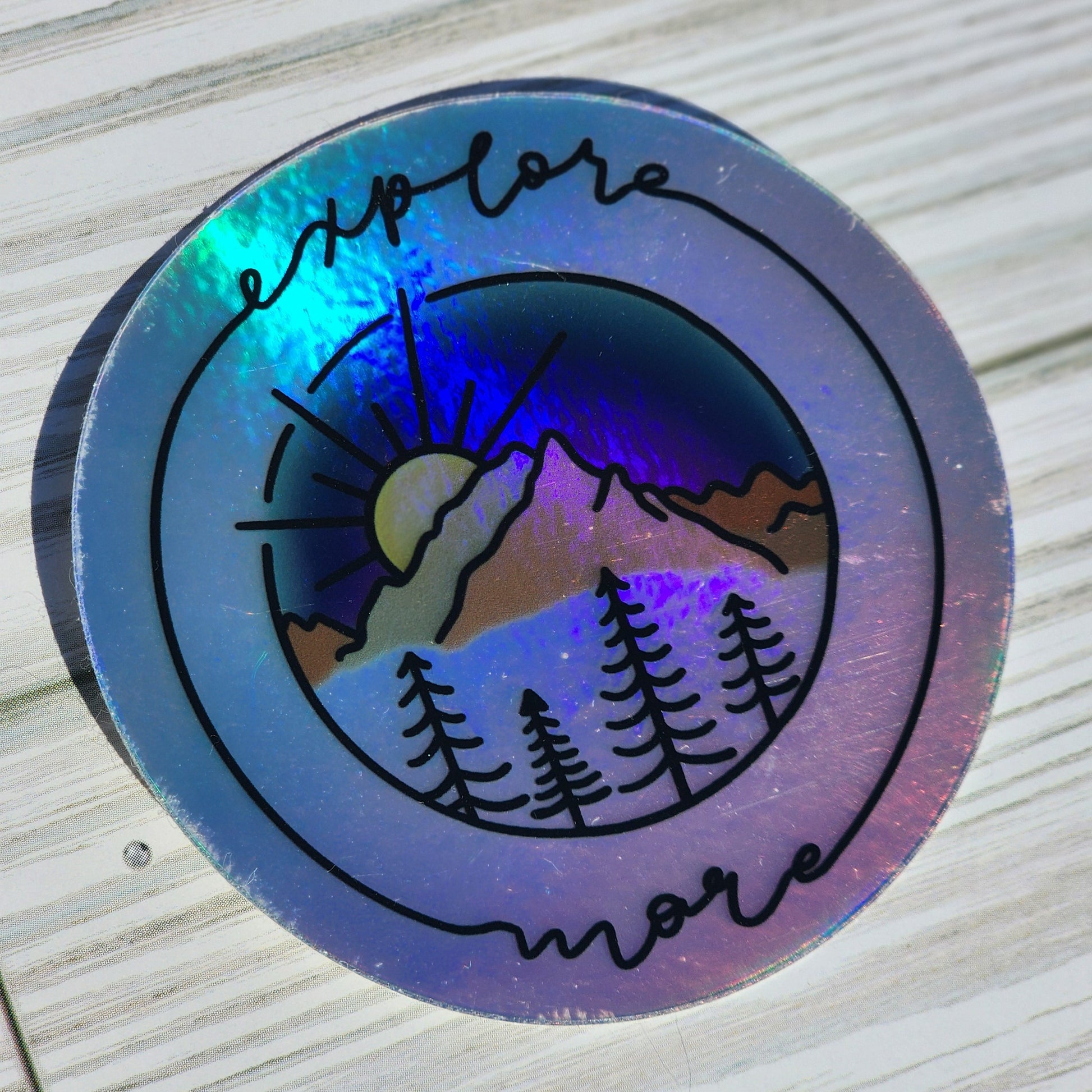 Vibrant mountain scape sticker with blue purple sunset, framed with cursive 'Explore More' on a holographic sticker