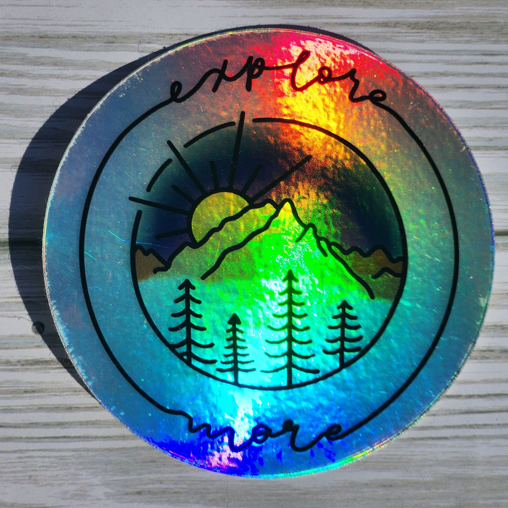 Vibrant mountain scape sticker with blue purple sunset, framed with cursive 'Explore More' on a holographic sticker