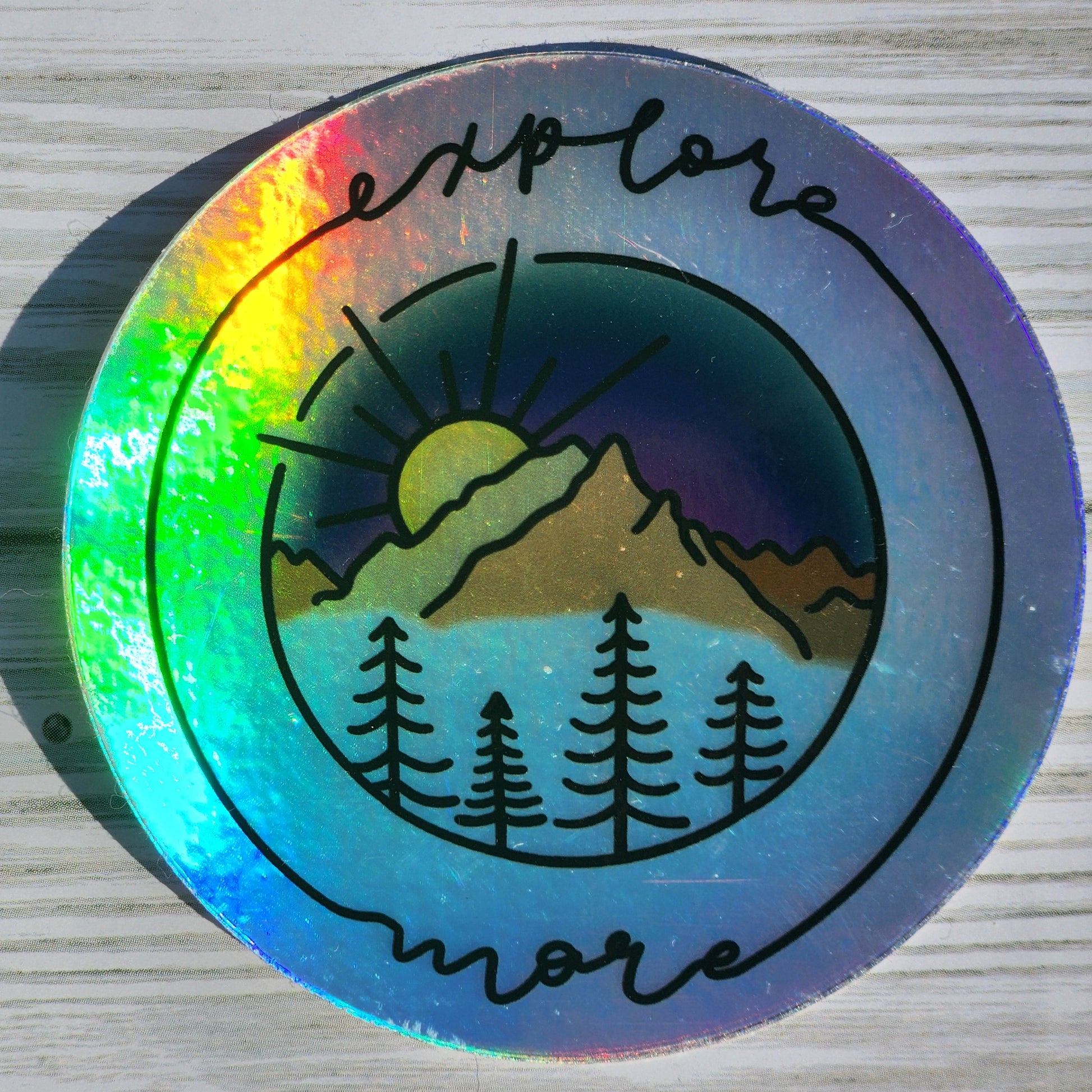 Vibrant mountain scape sticker with blue purple sunset, framed with cursive 'Explore More' on a holographic sticker