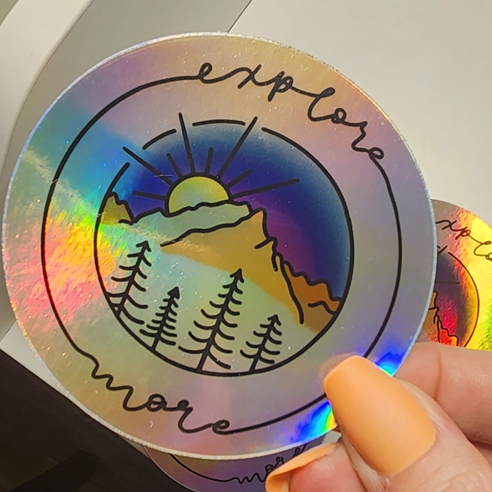Vibrant mountain scape sticker with blue purple sunset, framed with cursive 'Explore More' on a holographic sticker