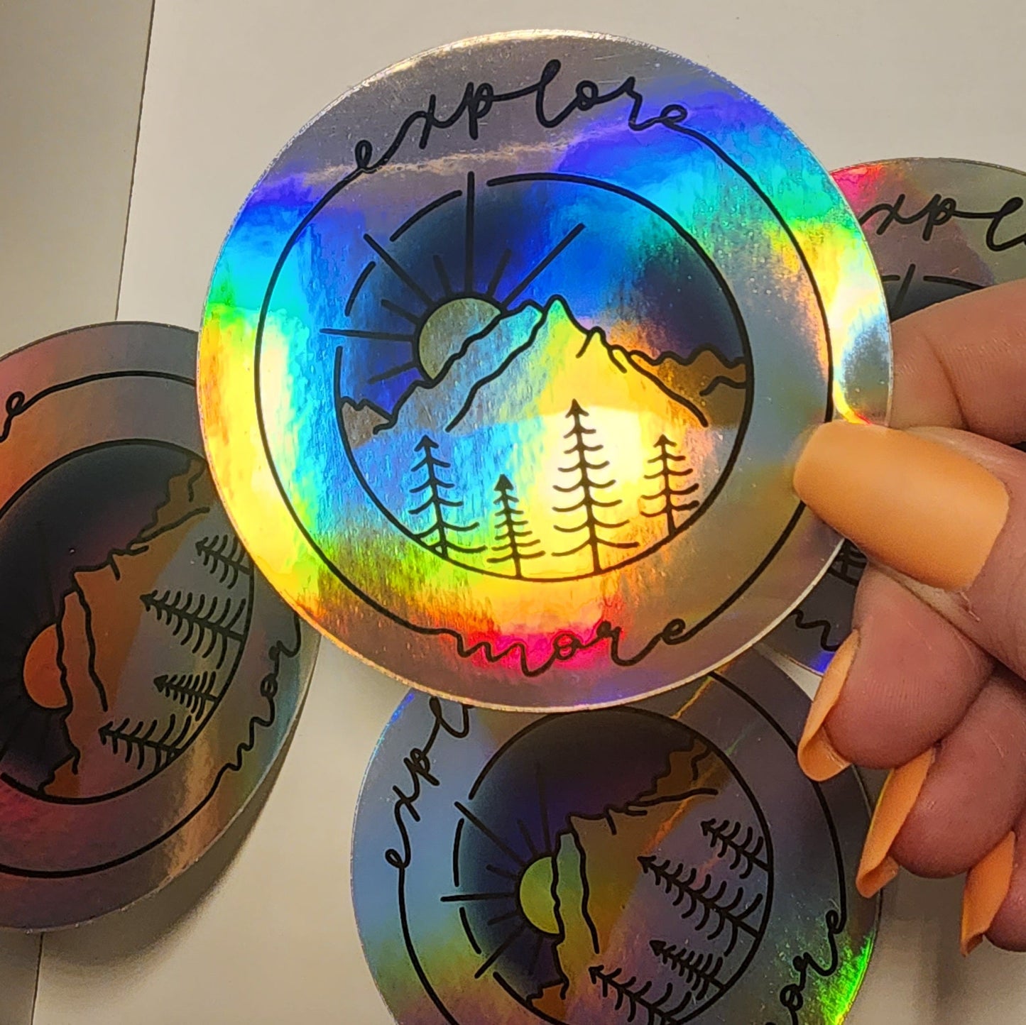 Vibrant mountain scape sticker with blue purple sunset, framed with cursive 'Explore More' on a holographic sticker showing reflection