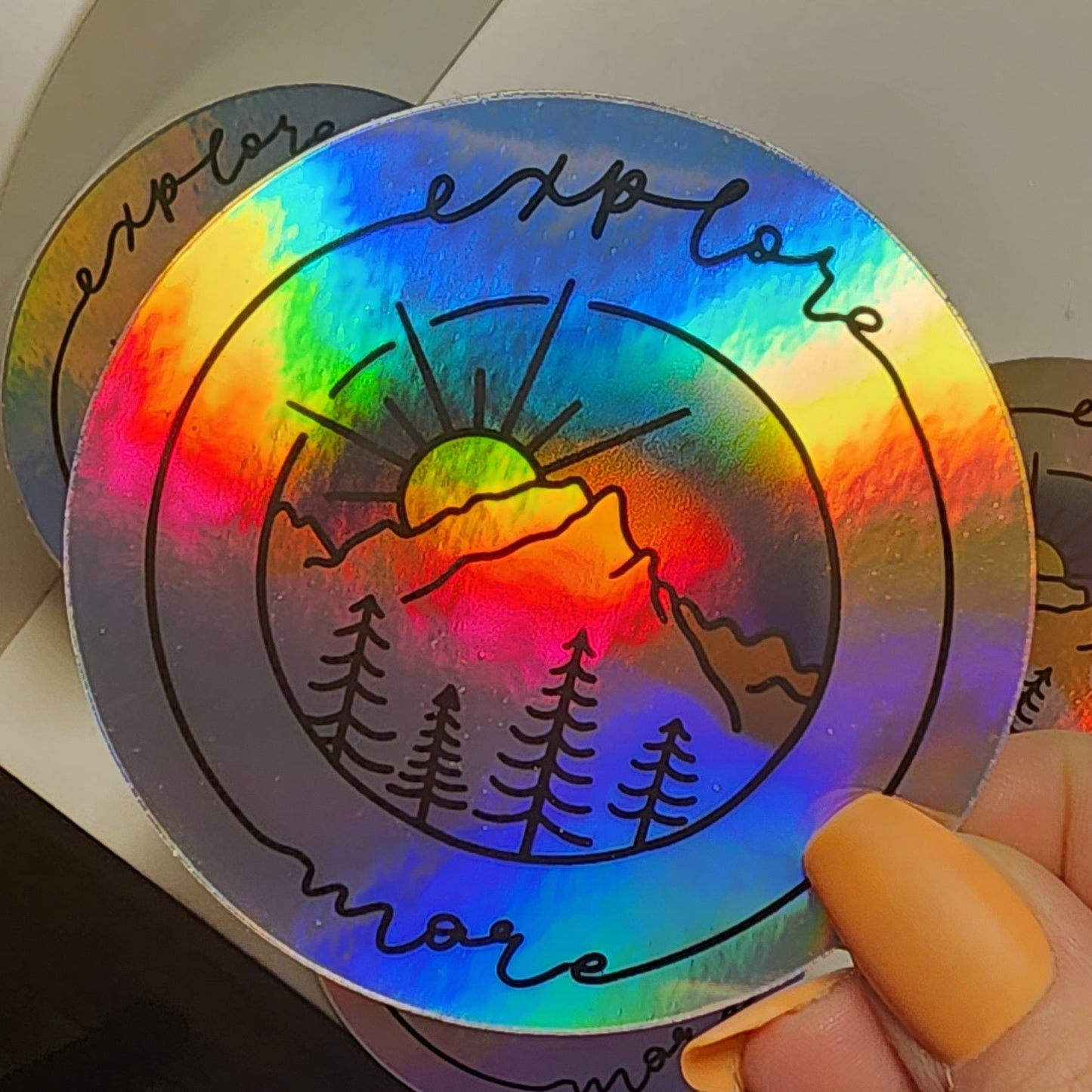 Vibrant mountain scape sticker with blue purple sunset, framed with cursive 'Explore More' on a holographic sticker