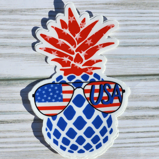Fourth of July-inspired sticker of a blue pineapple with red, star-covered crown and patriotic aviator sunglasses – one lens says 'USA', the other shows the flag.